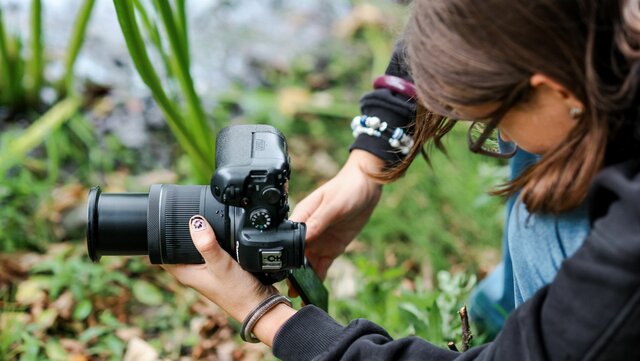 Frau mit Kamera fotografiert im Grünen mit Wasser im Hintergrund